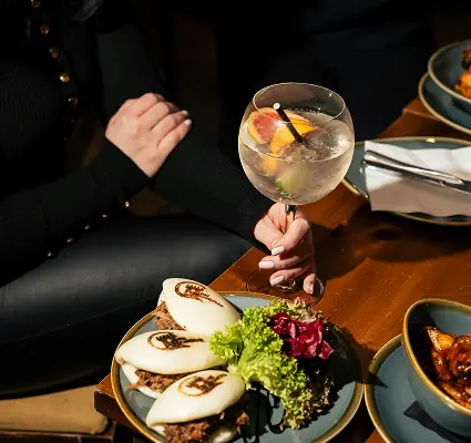 Person holding a cocktail above a table of bao buns and shared dishes